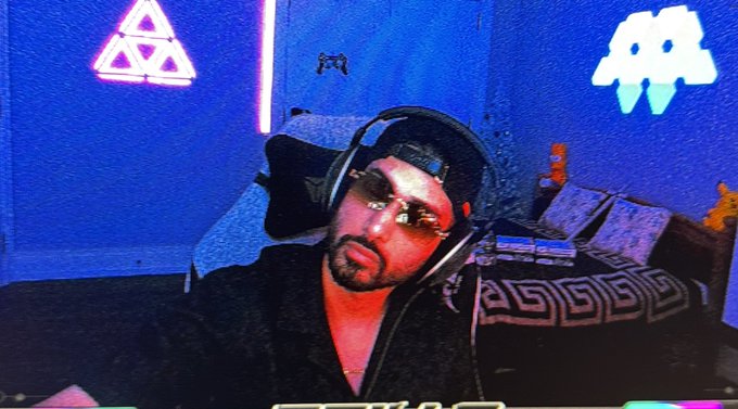 A man with sunglasses and a headset sits in a room illuminated by blue and pink lighting, with neon signs of a pyramid/triangle and a ghost in the background.