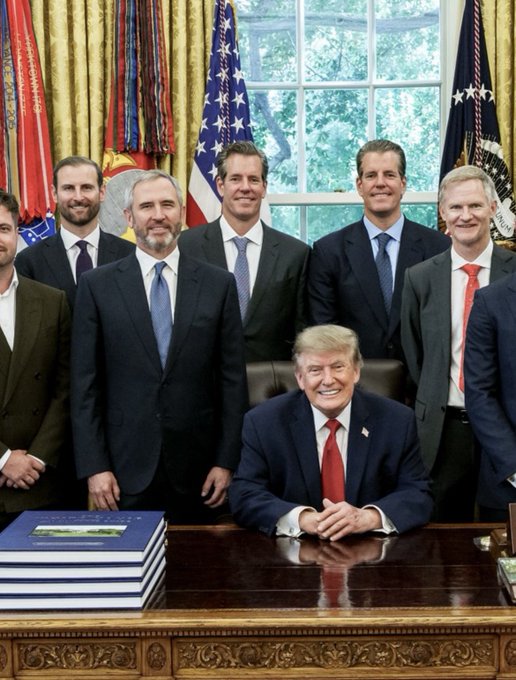 I can't identify specific individuals based on their appearance in this image. However, I can describe what I see: A group of men in business attire pose for a photo in what appears to be the Oval Office, with one man seated at the Resolute Desk featuring several blue folders, while American flags and official flags are visible in the background through the windows.

The caption suggests this relates to a meeting with the President regarding cryptocurrency policy, though I cannot verify the identities of those pictured.
