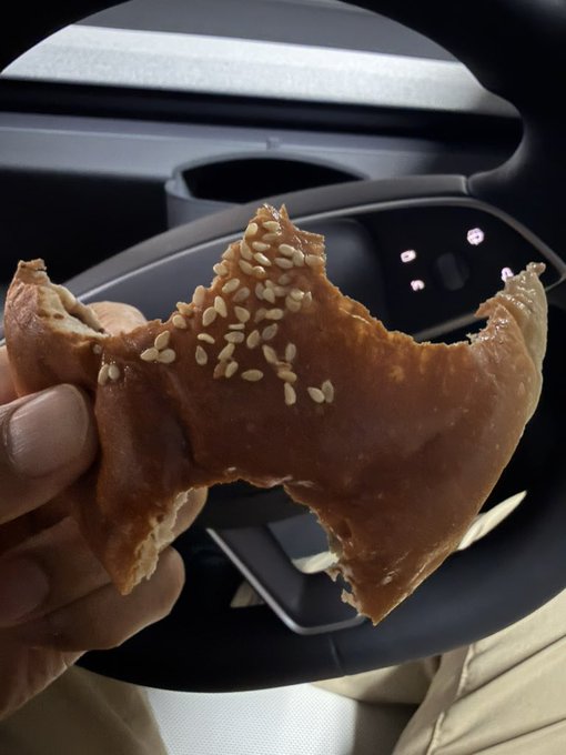 A partially eaten pastry topped with sesame seeds is held up inside a vehicle, with a steering wheel visible in the background.