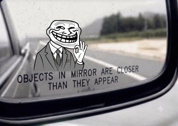 A photograph of a car's side mirror showing a road and landscape, with a Trollface meme character digitally added making an 'OK' hand gesture and the standard text 'OBJECTS IN MIRROR ARE CLOSER THAN THEY APPEAR'.