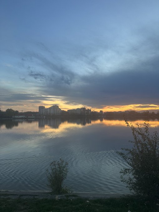 A scenic photograph captures a calm lake reflecting a sunset sky filled with dramatic clouds, with a city skyline visible on the distant shore.