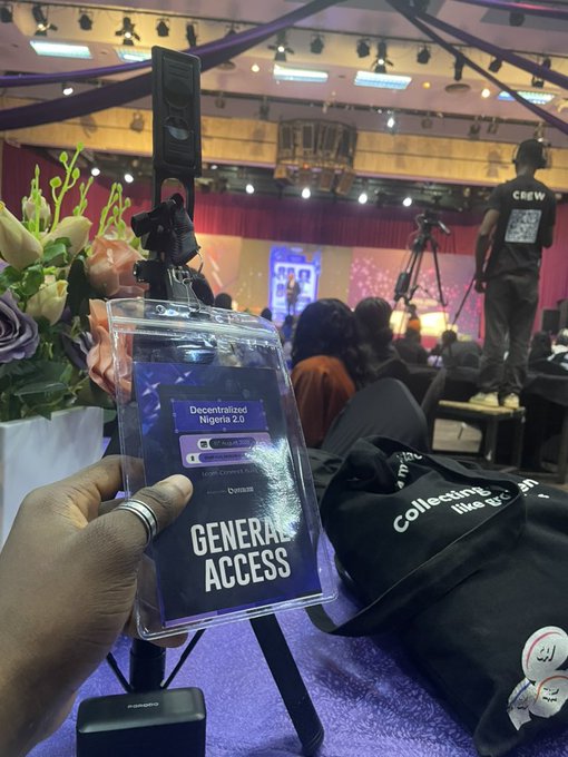 A person holds up a "General Access" badge reading "Decentralized Nigeria 2.0" at a conference event, with the scene showing a venue hall filled with attendees, crew members, and a bag branded "Collecting like a pro." The image captures the moment of attending the Decentralized Nigeria 2.0 event hosted by SuperteamNG, marking the badge as checked off the attendee's list.