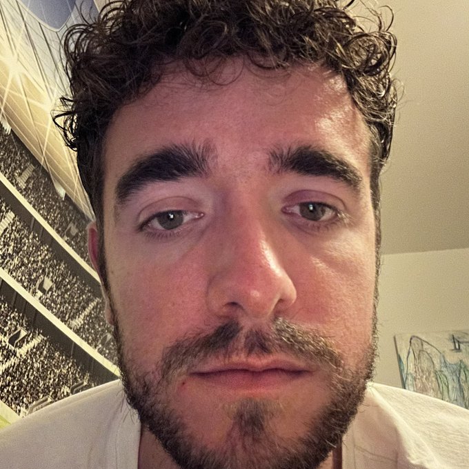 A man with curly dark hair, a beard, and visibly red, irritated eyes stares directly into the camera with a tired, defeated expression, appearing to be in a bedroom with posters visible behind him. The image visually captures the exhaustion and emotional toll of "Crypto Day 0," with his bloodshot eyes and weary look symbolizing the post-crash burnout implied by the caption "(It's Over)."