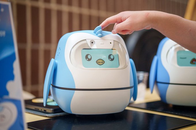 A person's hand reaches toward a small blue and white desktop robot with a cartoon penguin face on its screen, displayed on a table.