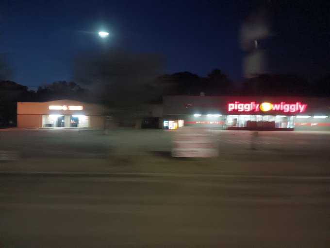 A blurry nighttime photograph taken from a moving vehicle, showing an illuminated "Piggly Wiggly" grocery store and another storefront with red neon lighting.