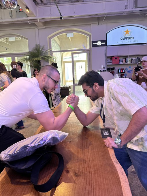 Two men lock hands in an intense arm wrestling match across a wooden table at an indoor venue, with a "SAPPORO LEGENDARY BIRU" sign and information booth visible in the background. A crowd watches the friendly competition, which humorously follows the poster's challenge of an arm wrestling rematch after their previous loss at "token 2049."