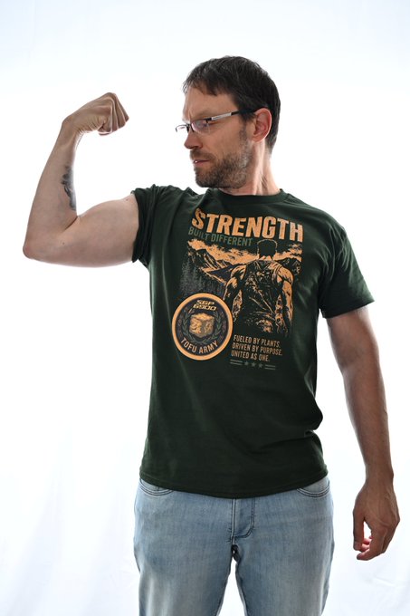 The image features a man wearing a green t-shirt with an orange circle design that says "SPX6900" on it, along with some text. He has his arm raised above his head, flexing his bicep muscles. The background is white, which makes the colors of the shirt and the design stand out more prominently. There are no other objects or people in the image, so the focus is solely on the man and his t-shirt.