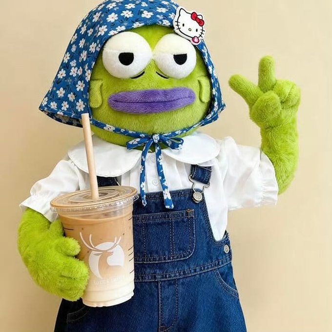 A green plush character wearing a blue floral headscarf, white blouse, and denim overalls holds an iced coffee cup and makes a peace sign.