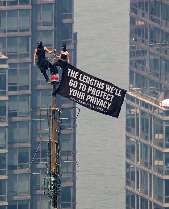 Two figures in black masks perch atop a cell tower between skyscrapers, unfurling a banner reading "THE LENGTHS WE'LL GO TO PROTECT YOUR PRIVACY — PROGRAMMABLE PRIVACY," visually embodying the post's point about companies going to extreme measures to safeguard user data.