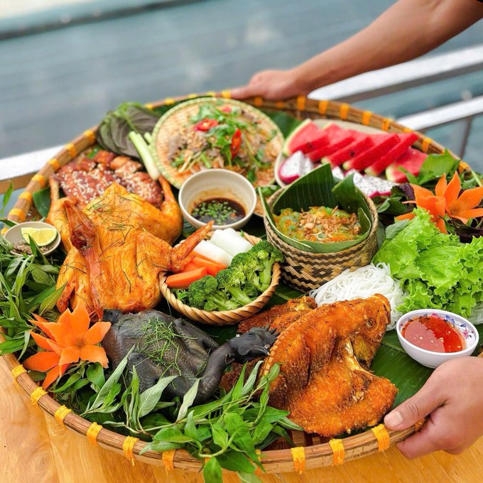 A large woven tray filled with a feast of foods including a whole roasted chicken, fried fish, fresh herbs, vegetables, and a papaya carving, presented on a boat.
