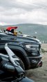 A rider in a red jacket sits atop a motorcycle on a mountain road, overlooking a valley with a small settlement nestled in the rolling green hills. The caption "闯入神的自留地！" ("Breaking into God's reserved land!") evokes a sense of adventure into a remote, untouched natural landscape.