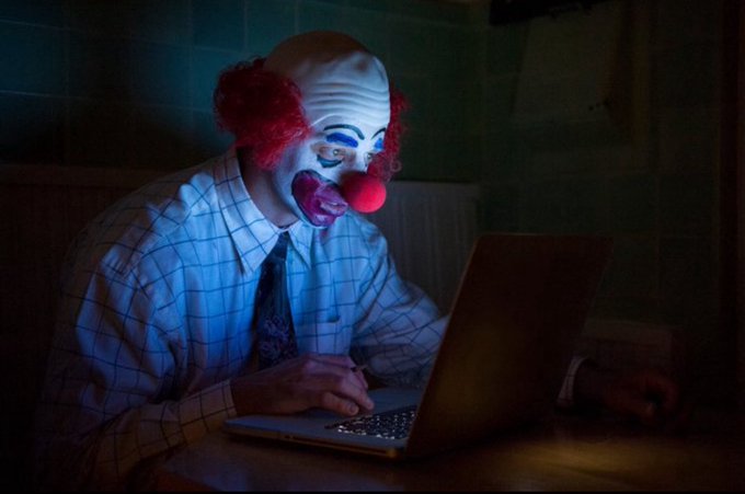 A person wearing a creepy clown mask and wig types on a laptop in a dimly lit room with blue lighting.