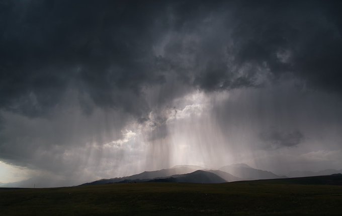 A dramatic landscape photograph showing dark, stormy clouds with bright crepuscular rays beaming down over a silhouetted mountain range and grassy plain.