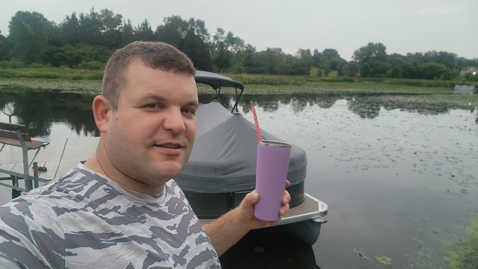 A man in a camo shirt holding a purple tumbler cup with a red straw, standing next to a pontoon boat on a calm lake surrounded by trees.