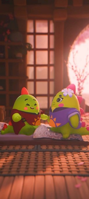 Two small, green, cartoonish characters with pink cheeks and red spikes sit together on a cushion in a warmly lit room with wooden floors and a window.