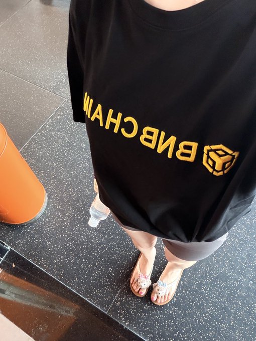 A person stands on a speckled floor wearing an oversized black t-shirt with yellow 'BNB CHAIN' text and a hexagonal logo, paired with flip-flops with floral decorations.