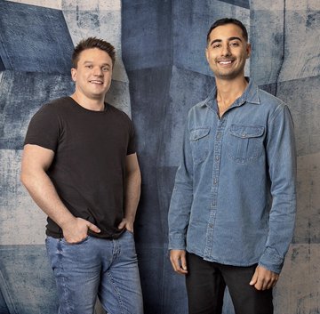 Ed Craven and Bijan Tehrani, co-founders of the crypto gambling site Primedice, pose together for a portrait against a blue-toned backdrop. The two men—one in a black t-shirt and the other in a denim button-up—smile at the camera, reflecting the friendly partnership described in the accompanying post about their journey from teenage RuneScape entrepreneurs to crypto gambling industry founders.