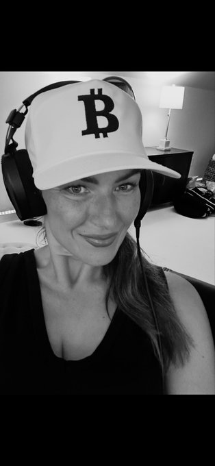 A black and white photo of a smiling woman wearing a white baseball cap featuring the Bitcoin (B) logo and large over-ear headphones.