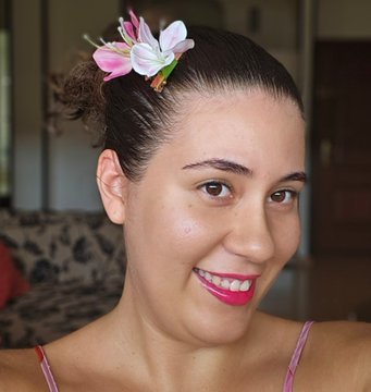 A smiling woman with dark hair pulled back, wearing a pink top and a pink and white flower tucked behind her ear.