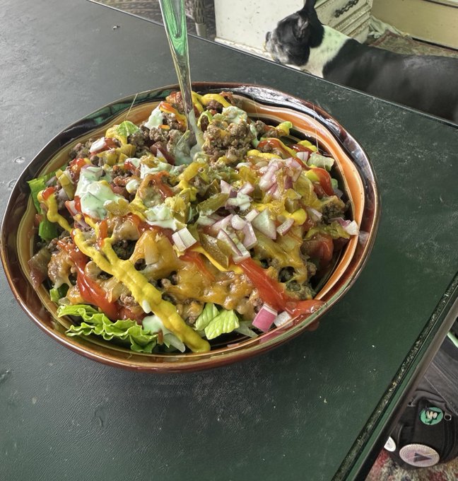 The image features a large bowl of food sitting on top of a table. The food appears to be a salad with various toppings, including lettuce, tomatoes, onions, and cheese. A dog can also be seen in the background, possibly observing the scene or waiting for its meal.