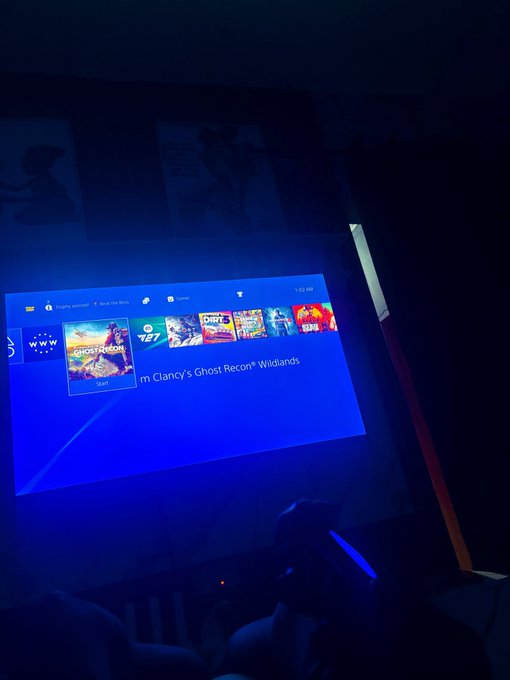 A dark game-room scene shows a TV displaying a console home menu with “Clancy’s Ghost Recon® Wildlands” selected, while silhouettes sit below the screen.