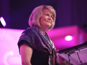 A woman with gray hair, wearing a patterned scarf and dark top, stands smiling at what appears to be a podium with stage lighting in the background, suggesting a speaking engagement or political event setting. The image likely depicts Senator Cynthia Lummis delivering remarks on her proposal for U.S. Bitcoin acquisition as a national debt reduction strategy.