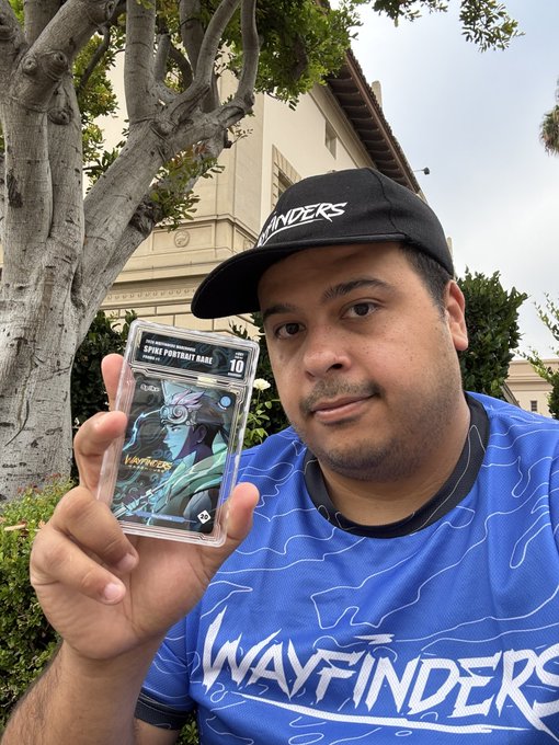 A man wearing a black 'Wayfinders' cap and blue jersey holds up a graded trading card featuring a 'Spine Portrait Rare' holographic artwork.