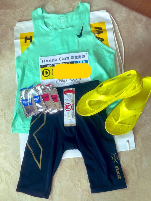 A flat lay of marathon gear laid out on a towel, including a mint Nike tank top with a "Honda Cars 南北北海道 Hakodate Marathon 2026" bib, dark compression shorts with "CR2" branding, bright yellow Nike racing shoes, two Amino Vital supplement packs, and an energy gel, all prepped for the runner's first full marathon at the Hakodate Marathon.