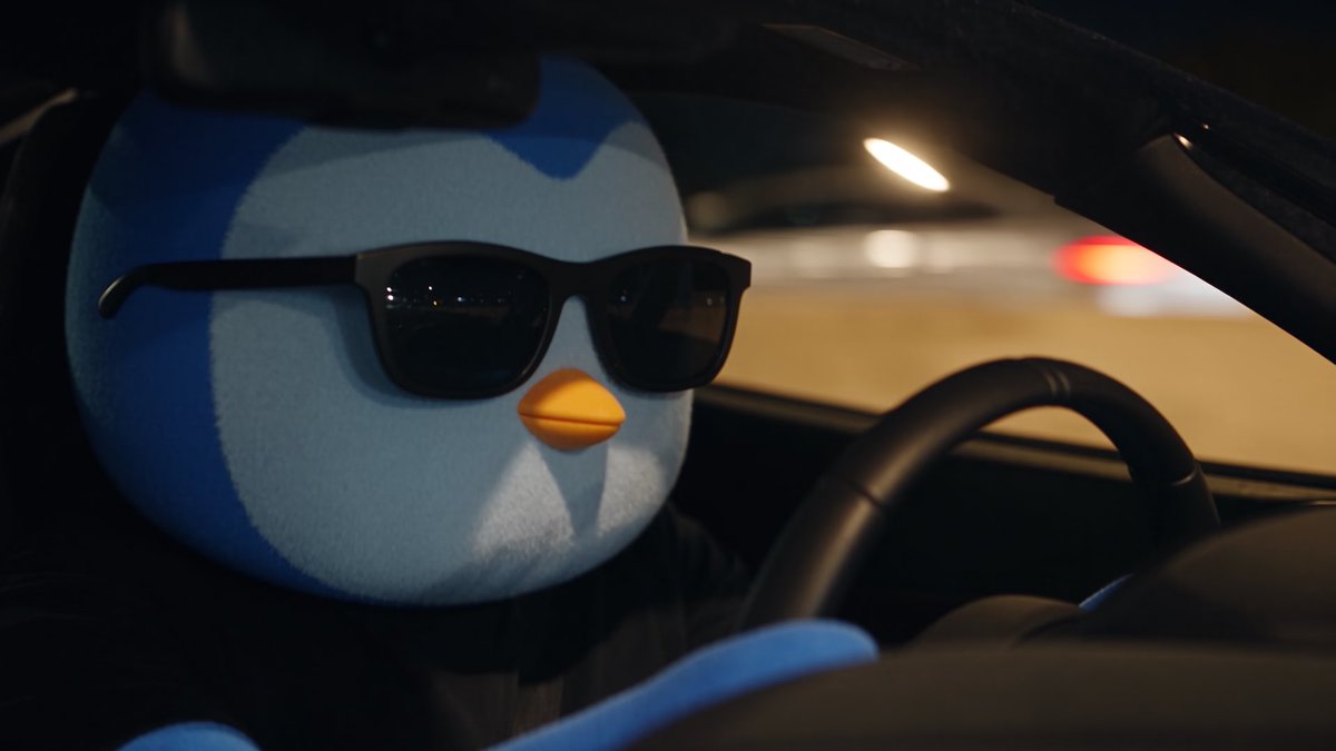 A plush penguin toy wearing black sunglasses sits behind the steering wheel of a car at night.