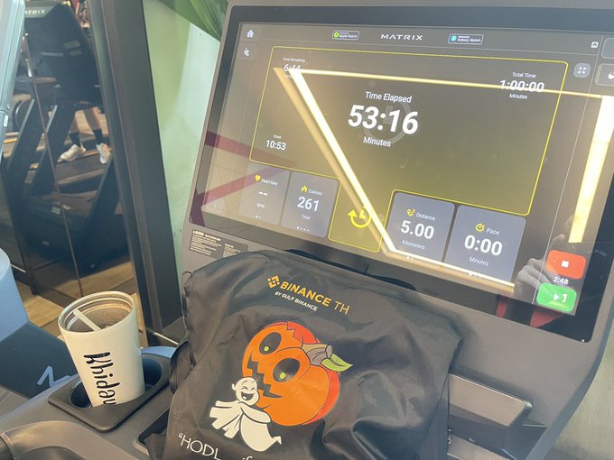 A Matrix brand gym treadmill screen shows a workout elapsed time of 53:16 minutes, with a Binance-branded drawstring bag and a Khadafi-branded cup resting on the console.