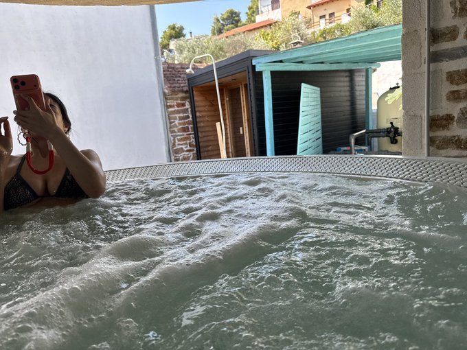 A photo of a woman in a black bikini in a bubbling hot tub, taking a selfie with a pink phone, with brick structures and greenery visible in the background.