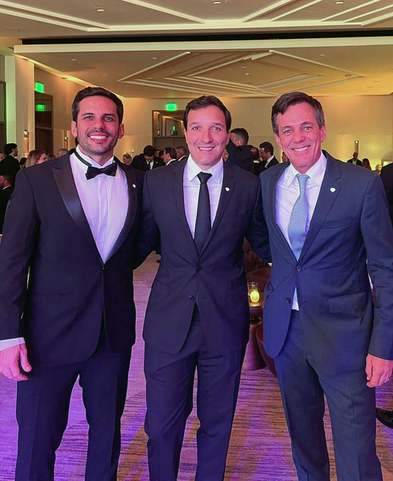 Three men in formal suits posing together at what appears to be a formal event or gala, smiling for the camera in a well-lit venue.