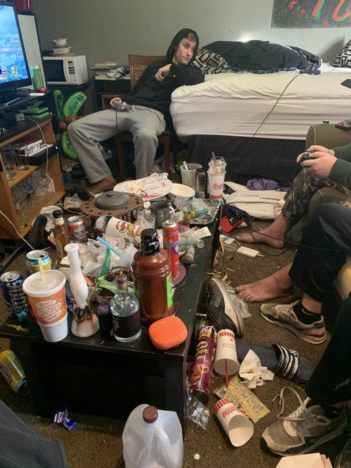 A cluttered dorm room with a person sitting on a chair making a peace sign, surrounded by a coffee table strewn with fast-food wrappers, soda cans, water bottles, and scattered shoes.