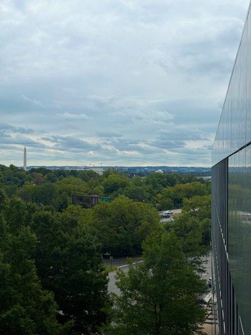 An elevated view shows lush green trees and a roadway, with the Washington Monument visible in the distant left. A modern glass building facade angles down the right side of the frame under an overcast sky.
