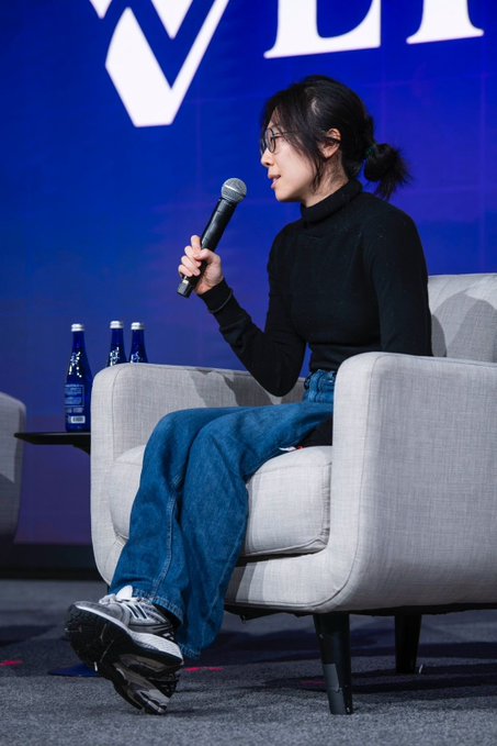 The image shows a woman sitting in an armchair, holding a microphone and speaking into it. She appears to be giving a speech or presentation. The background features a blue wall with a white "V" logo, which could possibly represent a company or organization related to finance or technology. There are three bottles placed on the table behind her, suggesting that she might have been discussing financial matters or sharing information about these products during her talk.