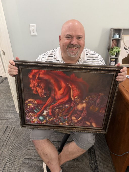 A bald man with a gray goatee smiling while holding a framed classical painting depicting a red apocalyptic scene with a horse and figures.