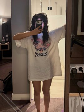 A person takes a mirror selfie in a hotel room wearing an oversized white "Rivals" graphic t-shirt, completely obscuring their face with a phone. The casual attire contrasts with the aggressive tone of the "FUCK KINGSMAN" caption, suggesting a humorous or rebellious mood unrelated to formal menswear.
