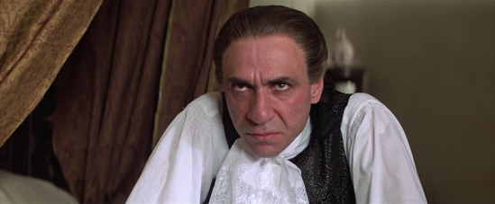 A scene featuring a man with slicked-back hair in period costume with a ruffled white shirt and vest, appearing to be from a historical drama film.
