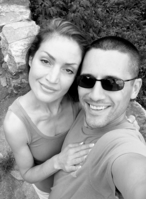 A black and white selfie of a smiling couple, with the woman in a tank top and the man wearing sunglasses, posed outdoors against a rocky, leafy background.