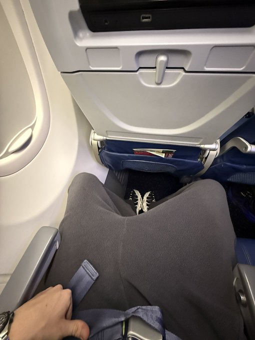 A passenger on a commercial flight to LA snaps a downward selfie showing their lap, feet, and a seatback pocket, humorously comparing their economy seat to a lavish private jet experience.