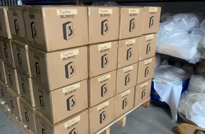 The image shows a large stack of boxes, with some of them labeled "Turbo Bloyz" and others labeled "Blockyz", indicating that they are related to a product or collection named Blockyz. The boxes appear to be made of cardboard and are neatly stacked on top of each other in a warehouse setting. The boxes have various sizes and shapes, suggesting that the collection may contain different types of products or items.