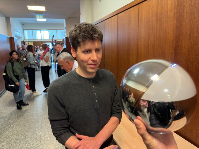A man with curly brown hair, wearing a gray henley shirt, stands smiling in a hallway while holding up a large reflective silver orb that mirrors the surrounding room and people. The post humorously suggests the glowing "orb" could serve as a government-subsidized gateway to free GPT usage, framing AI access as a mystical civic resource.