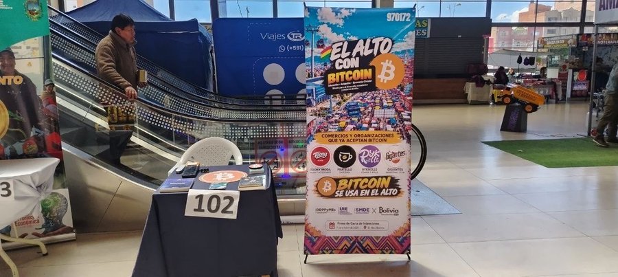 A promotional banner reading 'EL ALTO CON BITCOIN' (El Alto With Bitcoin) with the tagline 'BITCOIN SE USA EN EL ALTO' stands at booth 102 at an indoor event with escalators visible in the background.