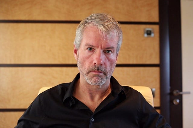 The image features a man wearing a black shirt who appears to be contemplating something or looking serious while sitting alone in a chair, possibly in an office setting near the camera. The photo captures his face and upper body at close range, with some of him visible in the background. There's no clear indication that he is engaged in any specific action related to the Bitcoin market crash and buying Bitcoins. It seems more like a casual moment or thought rather than active market trading.