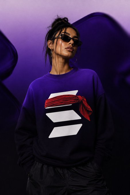 A woman in sunglasses and a purple-to-black sweatshirt stands against a purple backdrop; the shirt features a white geometric logo wrapped with a red cloth. The bold, energetic styling echoes the post’s “Solana speed and $GME” theme.