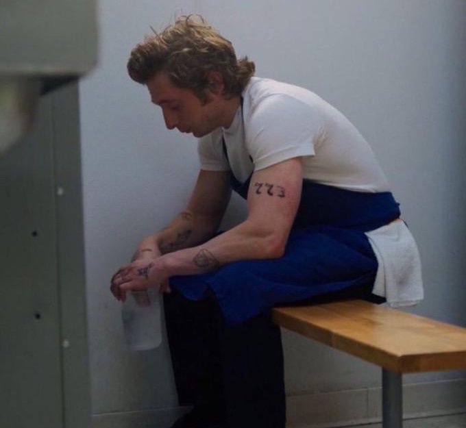 A young man with tattoos including "272" on his forearm sits hunched on a wooden bench in what appears to be a locker room or institutional setting, wearing a white t-shirt with a blue garment draped over his lap. The image evokes a sense of exhaustion and being overwhelmed, mirroring the post's caption where the person apologizes for being unable to respond to all requests while teasing the upcoming release of "novas mints."