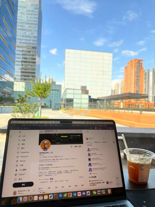 A laptop displays an X (formerly Twitter) profile for "@BTW0316" alongside an iced tea, set against a backdrop of modern skyscrapers under a blue sky—visually embodying the post's themes of lifelong learning, free markets, and intellectual freedom in a bustling urban workspace.
