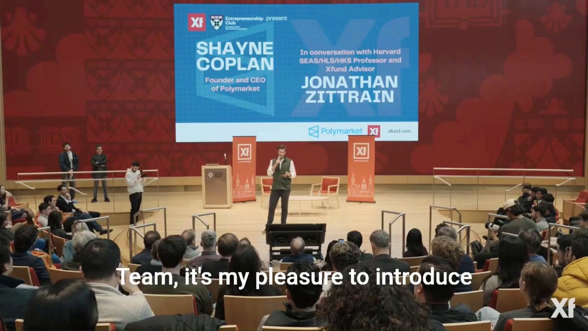 The image shows an event where Shayne Coplan, the founder of Polymarket, is giving a speech at Harvard University. The audience consists of people sitting in chairs while Shayne stands behind him, delivering his presentation. The setting appears to be indoors with a stage and a large screen displaying information about Shayne's company. The image also includes text that reads "Team, it's my pleasure to introduce you" and "Focus on what is in the image and how it relates to the post".