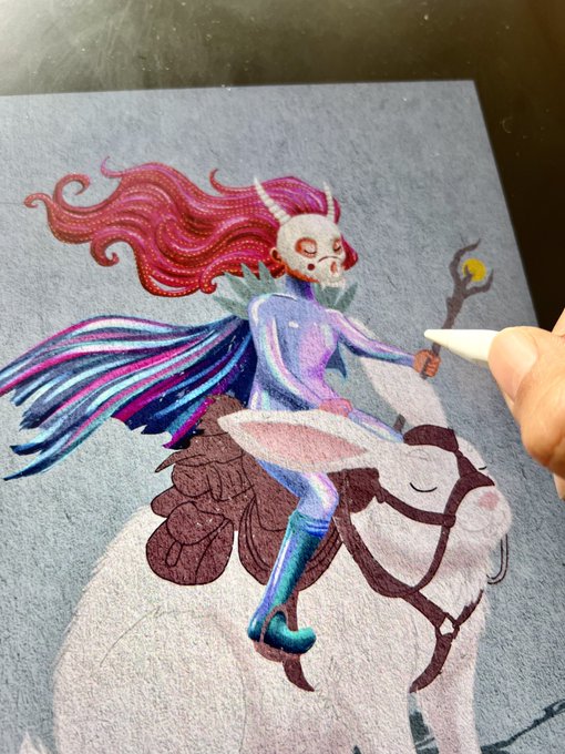 An artwork in progress depicts a horned, masked figure with flowing pink hair riding a white creature, rendered in colored pencil on gray paper as an artist's hand adds details.