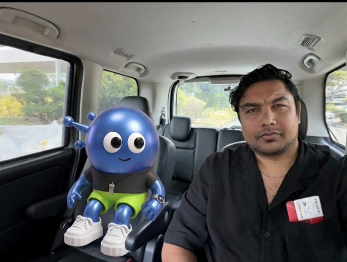 A man sits in the driver's seat of a car next to a blue cartoon robot character wearing a black shirt and green shorts.
